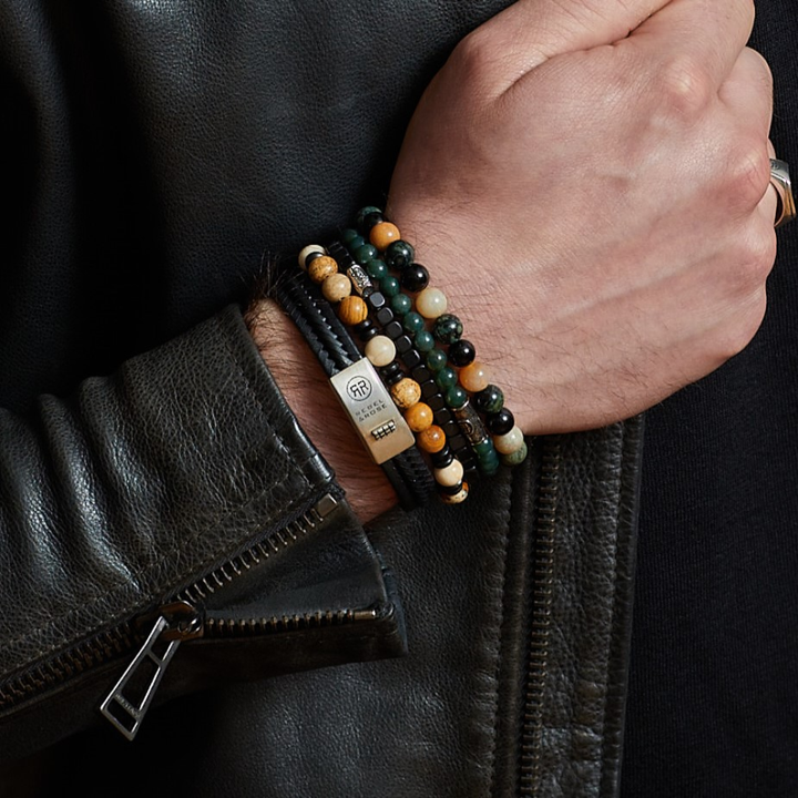 Een persoon in een donker leren jack laat verschillende kralenarmbanden zien, waaronder de Rebel & Rose kralen Woodstock - 8mm - Kralen Heren Armband in zwart, groen en bruin, samen met een metallic accent en een gouden ring.
