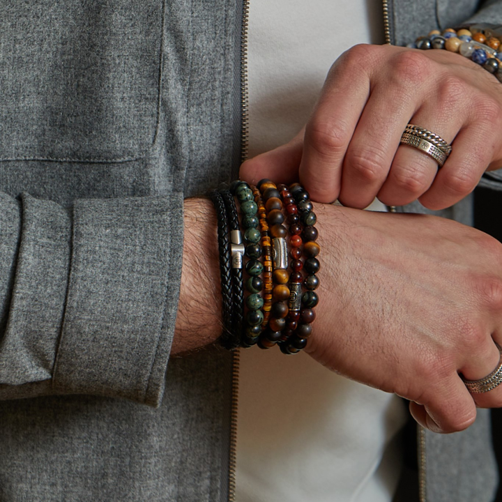 Een persoon in een grijs jasje en wit overhemd showt kralenarmbanden en ringen, waaronder een luxe Rebel & Rose Stormy Weather Vintage - 8mm Heren Kralen Armband, met kleurrijke kralen en leer op hun handen en polsen.