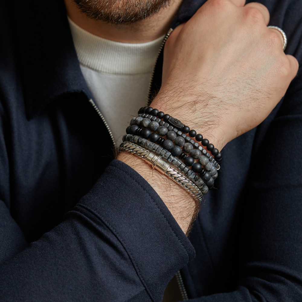 Een close-up van een pols en gezicht, gestyled met de Rebel & Rose Slices - Grey Seduction - 6mm Heren Kralen Armband, armbanden met zwarte kralen en zilveren kettingen, een zilveren ring, donkere jas met rits en witte coltrui voor een luxe sieradenlook.