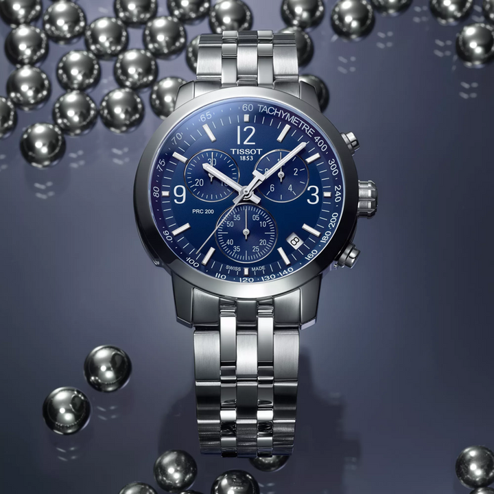 Een Tissot PRC 200 Chronograph herenhorloge met een blauwe wijzerplaat en zilverkleurige metalen band wordt getoond op een reflecterend oppervlak, omringd door metalen bollen.