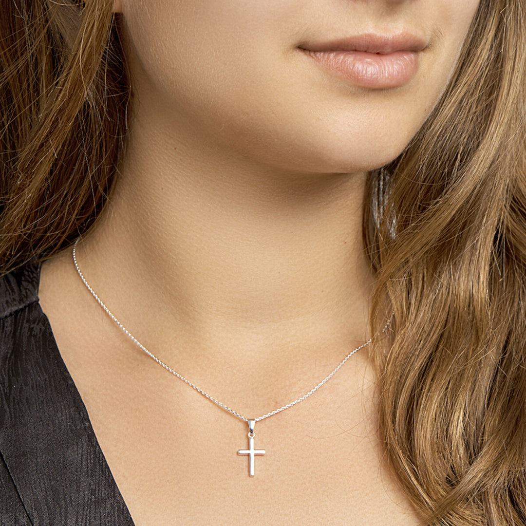 Een vrouw met lichtbruin haar draagt het Van der Kooij Collectie collier kruis 41 + 4 cm zilver wit, een fijne zilveren ketting met een klein kruisje. Gekleed in een top met donkere structuur zijn alleen haar lippen en kin gedeeltelijk zichtbaar voor een ingetogen look.