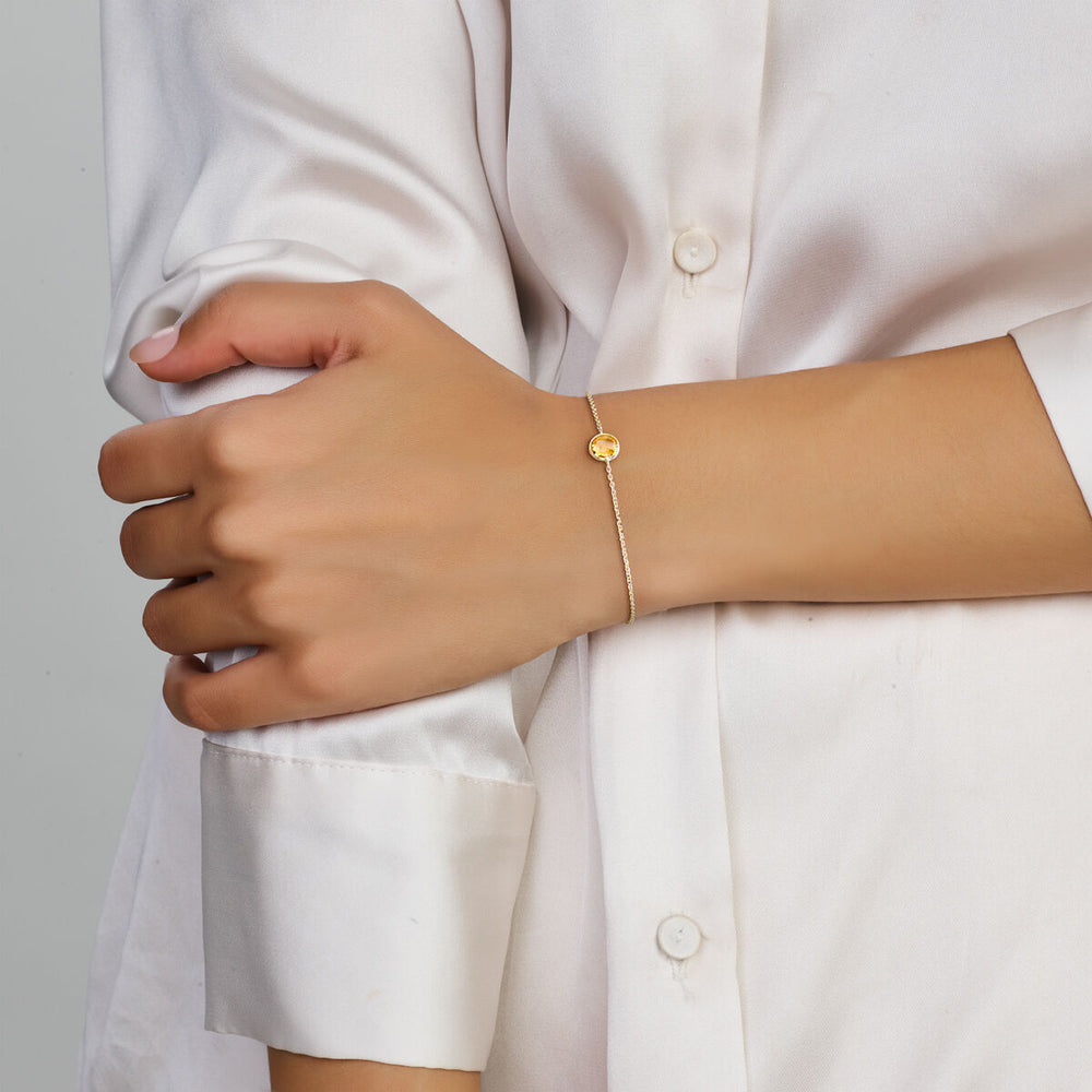 Een persoon in een wit overhemd met lange mouwen en knoopsluiting draagt de Van der Kooij Collectie Gouden armband dames citrien 14K, met een delicate ronde citrien bedel. De rechterhand houdt voorzichtig de linkerarm vast.