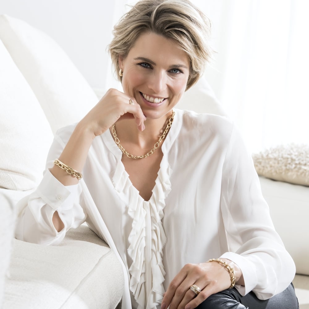 Een vrouw met kort blond haar en een witte blouse zit op een lichte bank, glimlacht en draagt de Geelgouden Armband 14 Karaat van Excellent Jewelry (20 cm, 10 mm), die tijdloze elegantie uitstraalt in een lichte, moderne woonkamer. Gratis verzending.