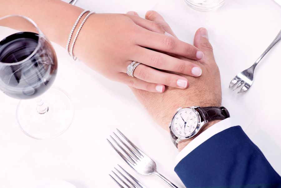 Close-up van een paar dat elkaars hand vasthoudt op een wit tafelkleed, met rode wijn en bestek; de vrouw draagt de Excellent Jewelry Witgouden Armband met Diamanten - 2.01 crt Briljant, 14 karaat, terwijl de man een blauw pak draagt.