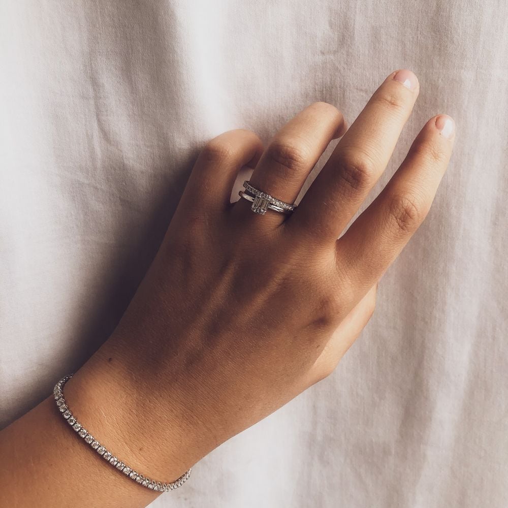 Een hand met een lichte huidskleur rust op witte stof, draagt de Excellent Jewelry witgouden tennisarmband met 63 briljanten - 3.01 crt diamant, 14 karaat, samen met twee zilveren ringen waaronder een elegant kruisontwerp bezet met kleine diamanten.
