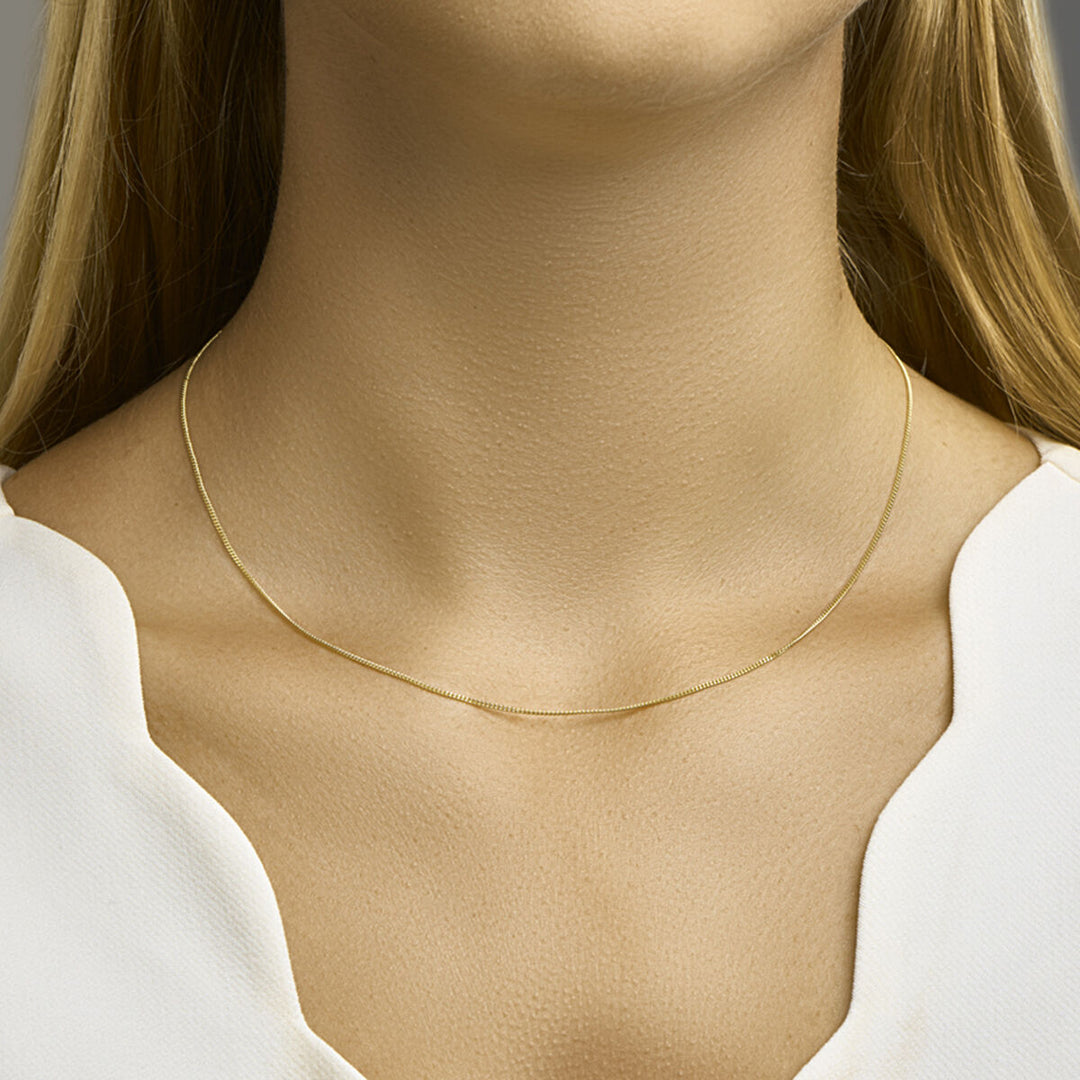 Een close-up van de hals van een vrouw met het Van der Kooij Collectie collier gourmette 1,0 mm 14K geelgoud. Ze draagt een wit topje met een ingekeepte hals, haar blonde haar is gedeeltelijk zichtbaar.