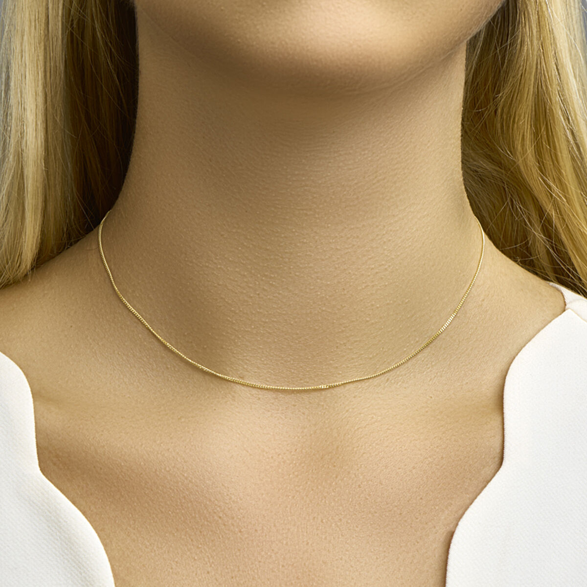 Een vrouw met blond haar draagt het Van der Kooij Collectie collier gourmette 1,0 mm 14K geelgoud om haar hals. Ze draagt een wit topje met V-hals tegen een neutrale achtergrond.