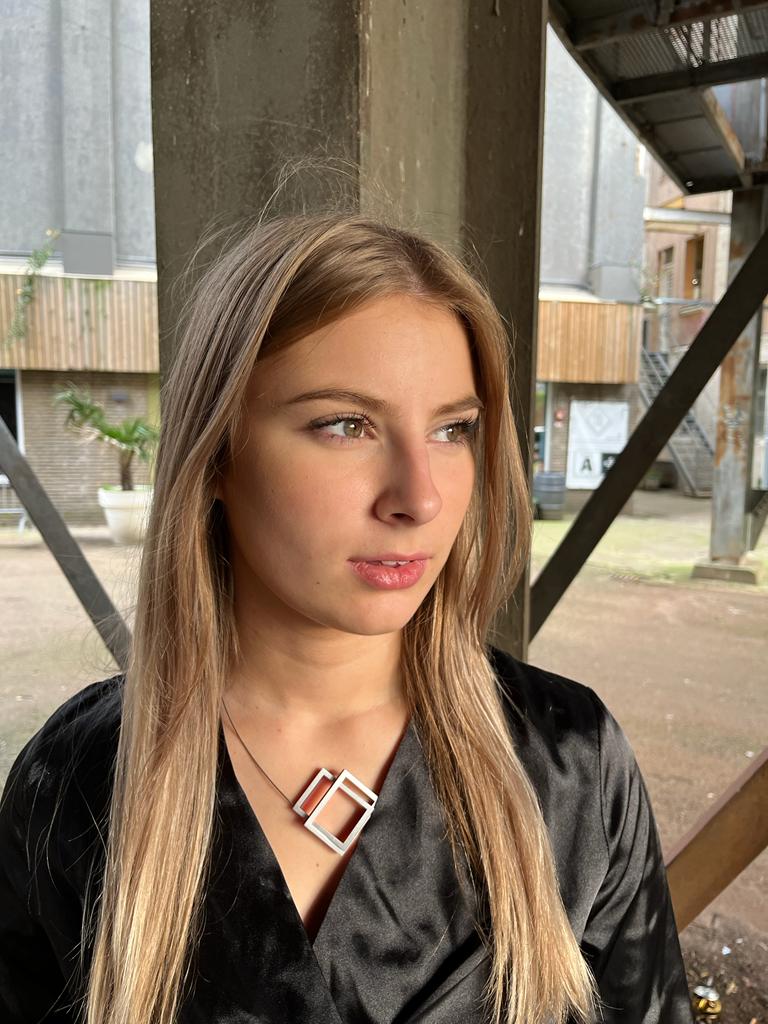 Een vrouw met lang blond haar staat onder een industriële constructie en kijkt opzij. Ze draagt een zwarte jas en de CLIC Creations Collier Twee rechthoeken in elkaar Zwart C30Z ketting, uniek geometrisch. De metalen balken en de moderne gevel van het gebouw vangen stedelijke elegantie.