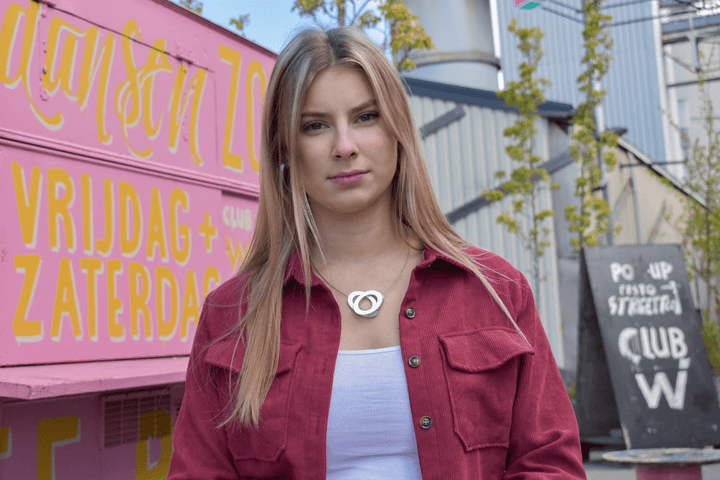 Een persoon met lang blond haar staat voor een roze bord met gele tekst, draagt een rood shirt en een wit topje. Op de achtergrond staat een krijtbord met de tekst Pop Up. Het lijkt erop dat de setting buiten is en ze dragen de Collier Ovalen op elkaar Goud|Geel C70G ketting van CLIC Creations.