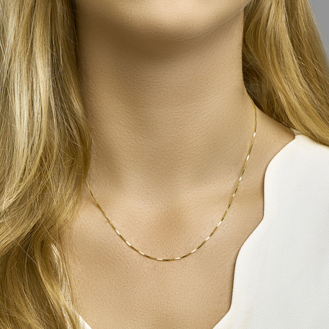 Een vrouw met lang blond haar draagt een Van der Kooij Collectie Venetiaans collier 0,8 mm 14K geelgoud, gecombineerd met een wit topje met inkeping. De focus ligt op de ketting, die haar nek en schouders accentueert.
