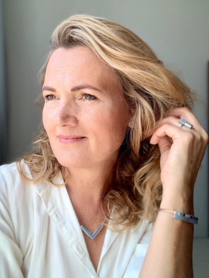 Een vrouw met golvend blond haar kijkt peinzend opzij, gekleed in een wit shirt en minimalistische sieraden: een handgemaakte driehoekige ketting van CLIC Creations uit Nederland en de veelzijdige Ring Vierkanten R6. Haar uitdrukking is kalm en beschouwend.