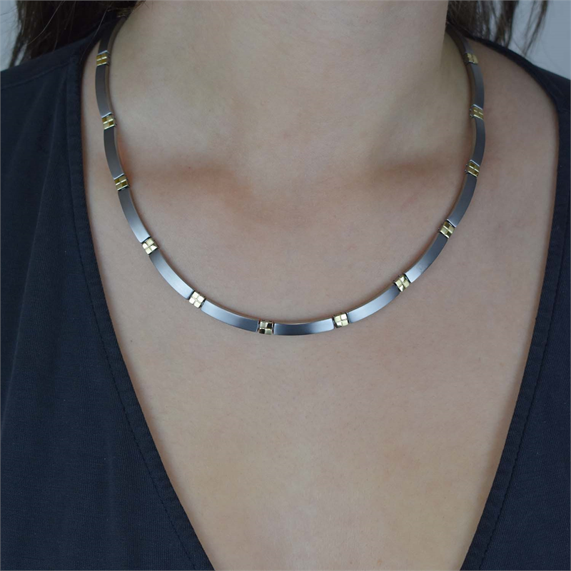 Een close-up toont een persoon die de Boccia Titanium Collier Bicolor 0831-02 draagt - een platte, zilveren en gouden halsketting van Boccia - die soepel langs het sleutelbeen rust tegen een donker topje met v-hals.