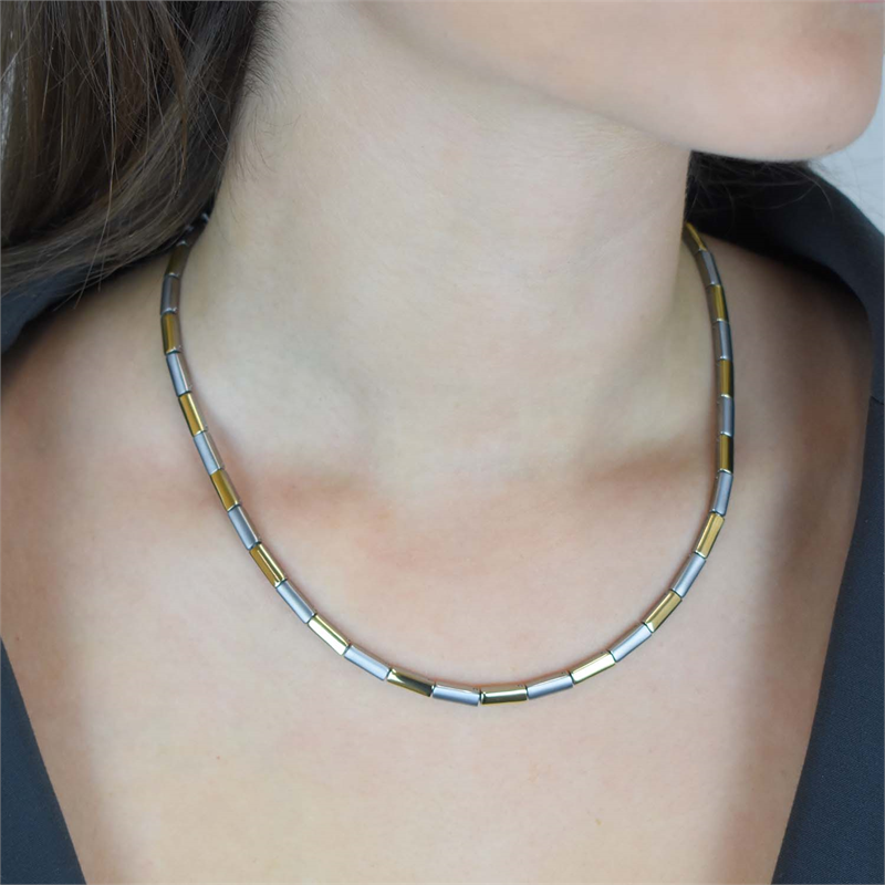 Een vrouw draagt de Boccia Titanium Collier Bicolor 0866-02-een korte halsketting van Boccia met afwisselend gouden en zilveren rechthoekige kralen, afgebeeld tegen haar blote nek en sleutelbeen, een deel van een donker topje zichtbaar.