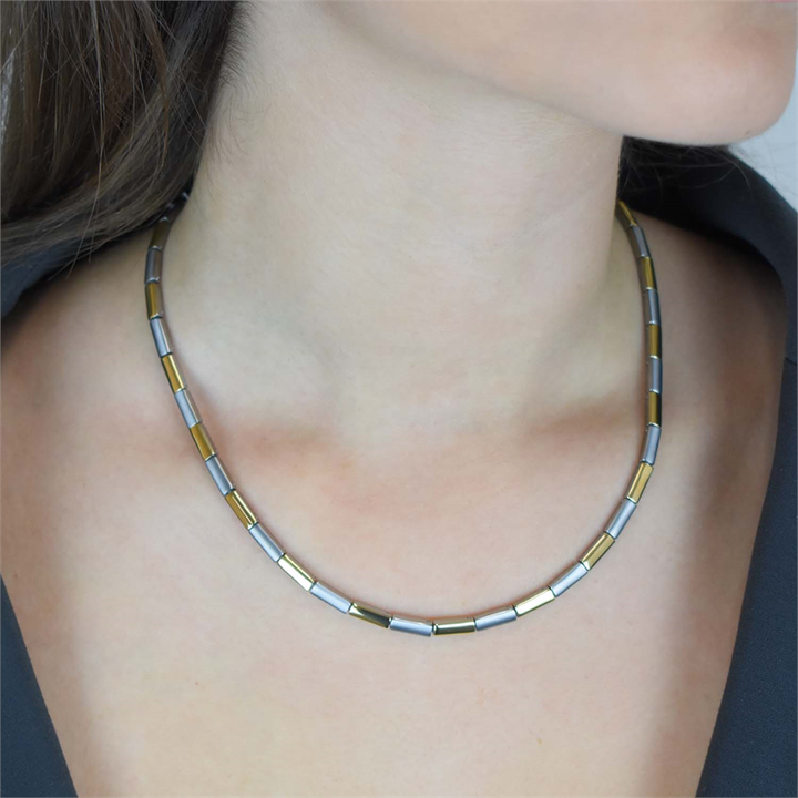 Een vrouw draagt de Boccia Titanium Collier Bicolor 0866-02-een korte halsketting van Boccia met afwisselend gouden en zilveren rechthoekige kralen, afgebeeld tegen haar blote nek en sleutelbeen, een deel van een donker topje zichtbaar.