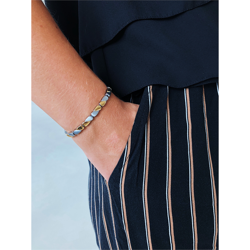 Een persoon in een zwart topje en een zwarte broek met beige verticale strepen, SKU 03016-02, poseert met één hand in zijn zak en toont de Boccia Titanium Armband Bicolor van Boccia om zijn pols.
