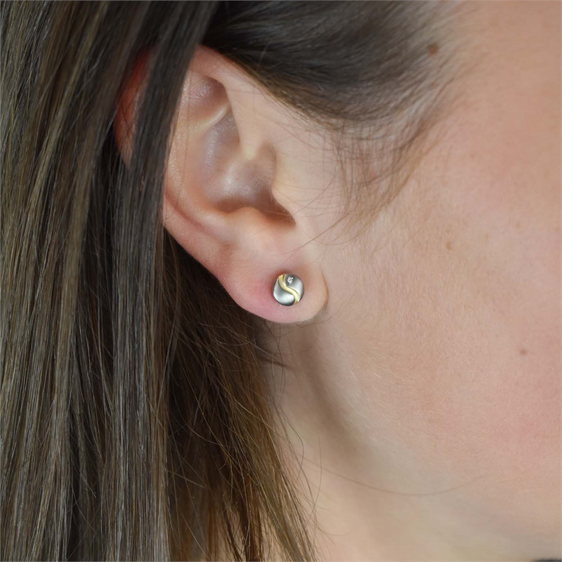 Close-up van een oor met Boccia Titanium Oorst. Tweekleurige oorsteker met een diameter van 2/0,01 - 05028-04 van Boccia, met een klein zilver-gouden wervelpatroon. Bruin haar omlijst het oor, met zichtbare huiddetails.