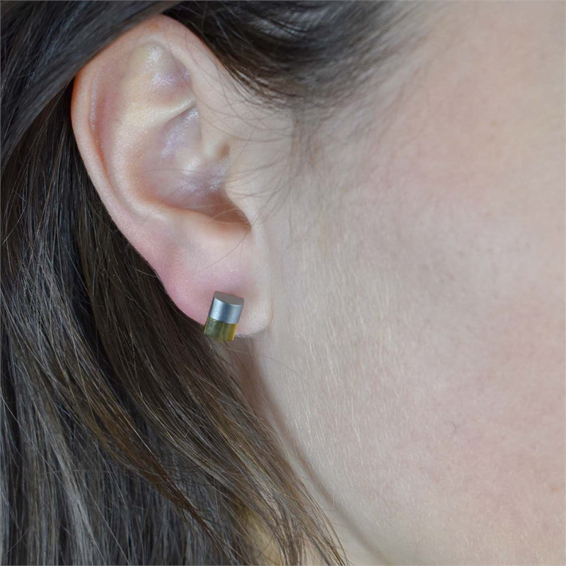 Close-up van het oor van een persoon met Boccia Titanium Oorstekers Bicolor 0509-03 oorstekers met een metallic grijze bovenkant en een doorschijnende groene onderkant. Deze moderne oorknoppen van Boccia zijn perfect als duurzame sieraden.