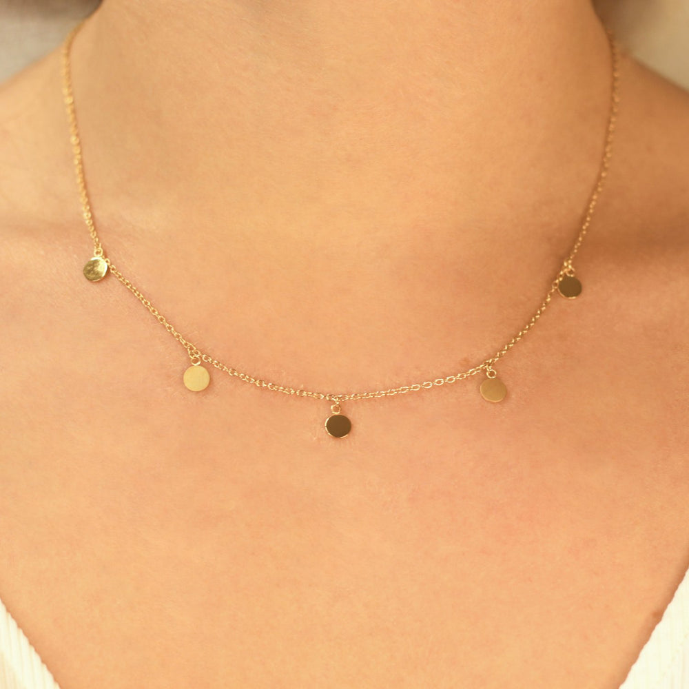 Een close-up van een persoon die de Boccia Titanium Collier gp 08064-02 van Boccia draagt, een delicate goudkleurige ketting met kleine, ronde, platte bedeltjes gelijkmatig verdeeld langs de ketting, die net boven het sleutelbeen rust.
