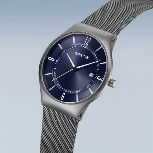 Het Bering 15940-378 herenhorloge heeft een blauwe wijzerplaat met witte uurmarkeringen, een datumvenster op 3 uur, saffierglas en een mesh metalen band. Het wordt weergegeven op een lichtblauwe achtergrond met kleurverloop.