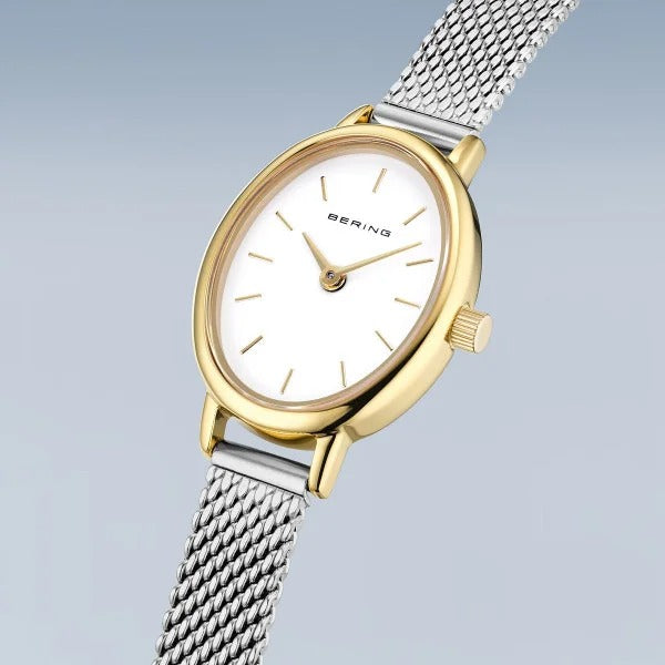 De Bering 11020-010 dameshorloge heeft een goudkleurige ronde kast, witte wijzerplaat met eenvoudige markeringen, quartz uurwerk, saffierglas en een zilverkleurige mesh band, afgebeeld tegen een lichtblauwe achtergrond.