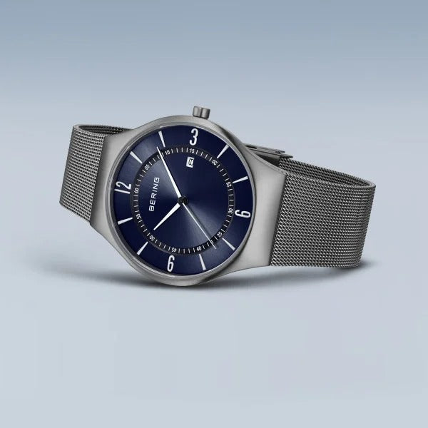 De Bering 15940-378 herenhorloge heeft een zilverkleurige mesh band, ronde donkerblauwe wijzerplaat met witte uurmarkeringen, saffierglas, datumvenster en zichtbare BERING branding. Het horloge wordt weergegeven op een lichtblauw oppervlak.