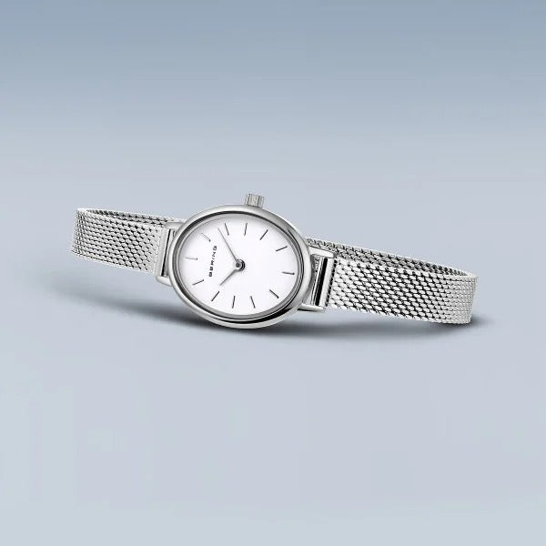 Het minimalistische Bering 11020-004 dameshorloge heeft een witte ovale wijzerplaat met slanke zwarte uurmarkeringen en een mesh band, prachtig tegen een lichtblauwe achtergrond.