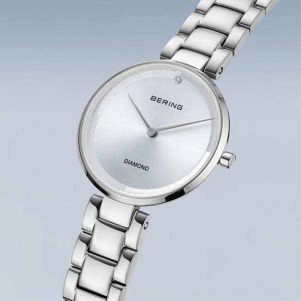 De Bering 17529-700 is een zilverkleurig dameshorloge met een metalen schakelband, minimalistische witte wijzerplaat, één diamant op 12 uur, saffierglas en alleen BERING en DIAMOND tekst op de wijzerplaat.
