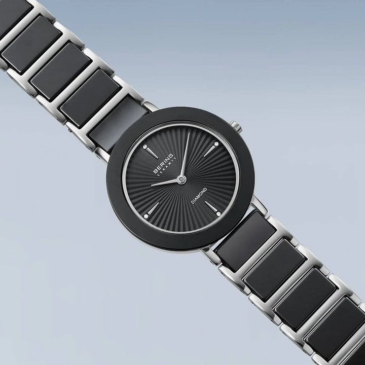 Het Bering dames horloge 11429-702 heeft een moderne zwarte en zilveren band, ronde zwarte wijzerplaat, minimalistische markers, "Diamond" detail op de wijzerplaat en saffierglas - alles tegen een zacht lichtblauw kleurverloop.