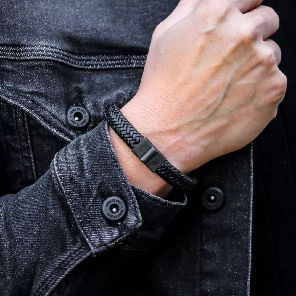 Een persoon in een zwart spijkerjack showt een Rebel & Rose Full Metal Herringbone Black Black armband om zijn gedeeltelijk gebalde pols in de buurt van de knopen van het jack.