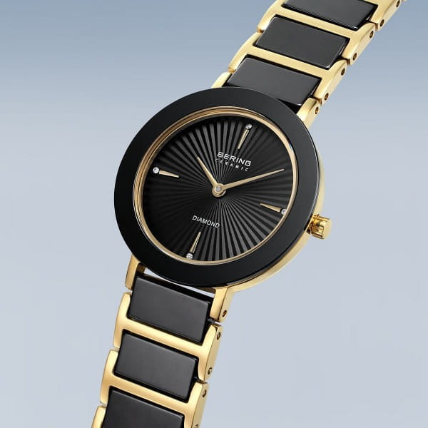 De Bering dames horloge zwarte wijzerplaat - 11429-732 van Bering heeft een zwarte en gouden band, zwarte ronde wijzerplaat, gouden uurmarkeringen en minimalistische diamanten details, weergegeven in een hoek tegen een lichtblauwe achtergrond.