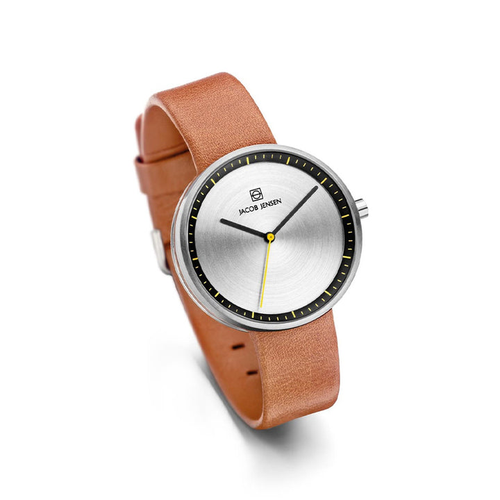 Een strak Jacob Jensen quartzhorloge, model 281, heeft een ronde zilveren wijzerplaat met minimale zwarte uurmarkeringen en een gele wijzer. Het heeft mineraalglasbescherming, een bruine leren band en het logo van het merk op de wijzerplaat.