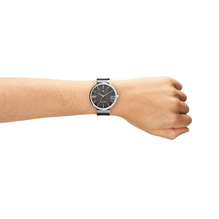 Op de arm is een strak Jacob Jensen 708 horloge te zien met een zwarte wijzerplaat en zilveren band, met eenvoudige uurmarkeringen, wijzers en een nauwkeurig quartz-uurwerk op een effen witte achtergrond.