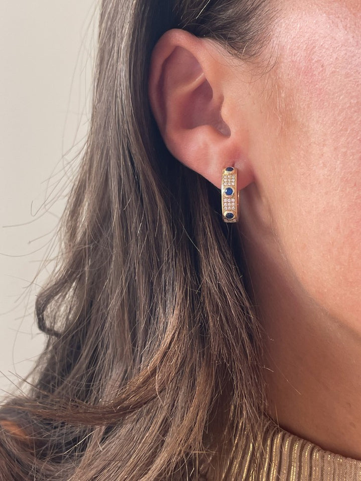 Een close-up van een vrouw met Excellent Jewelry's geelgouden collier met blauwe saffier (0.50 crt) en diamanten (0.24 crt), 14 karaat, met gouden hoepel oorbellen en bruin haar gedeeltelijk zichtbaar op de achtergrond.