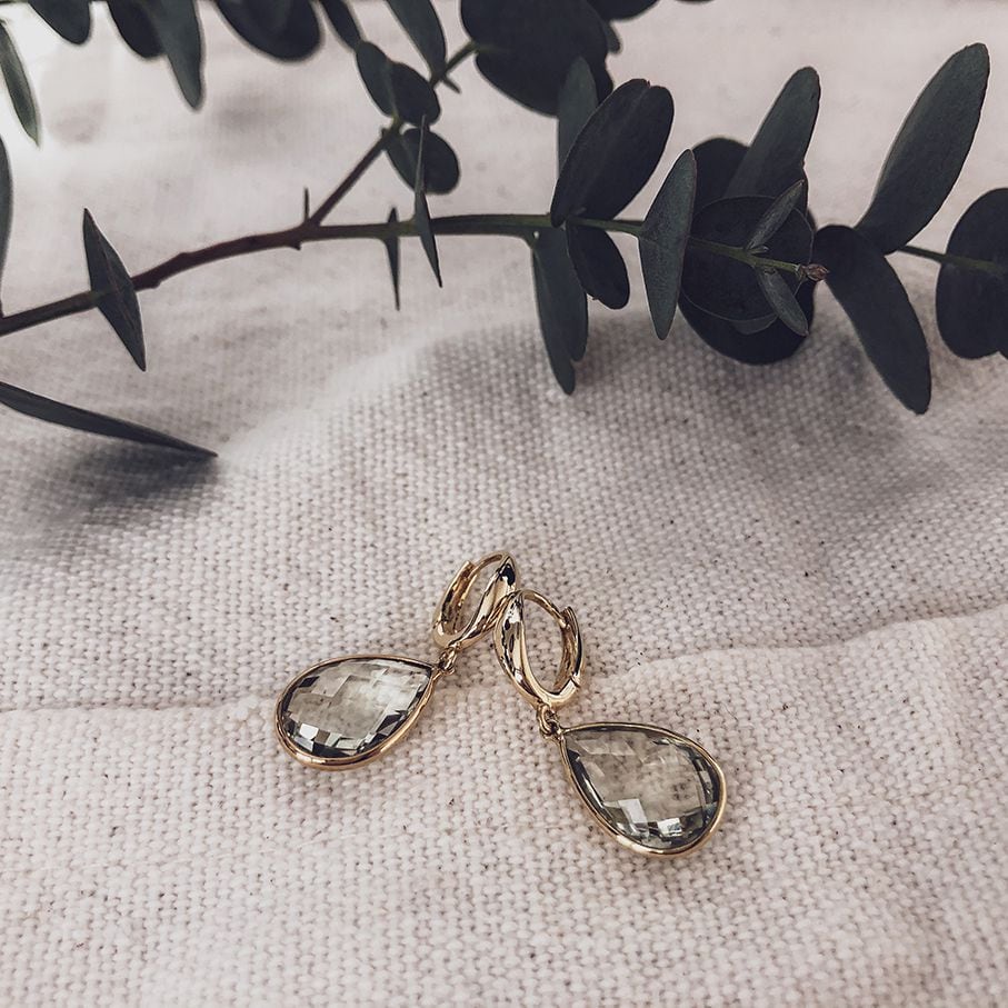 De Stralende Geelgouden Oorringen met Citrien Aanhangers van Excellent Jewelry liggen op textuur crème stof, met een eucalyptus tak diagonaal op de achtergrond.