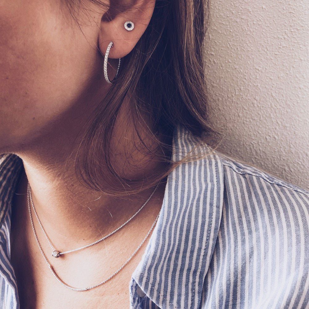 Close-up van een vrouw met Excellent Jewelry Bicolor Gouden Oorknoppen met Saffier en Diamanten 0,11ct, zilveren hoepel en stud oorbellen, twee delicate zilveren kettingen met kleine hangers, en een blauw-wit gestreept shirt. Haar haar bedekt gedeeltelijk haar oor.