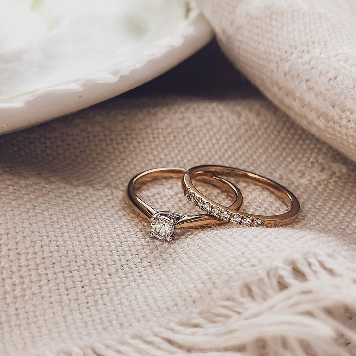 Twee Elegante Bicolor Gouden Ringen van Excellent Jewelry liggen op beige stof; één ring heeft een enkele diamant, de andere is bezet met briljanten van 0.19 crt.