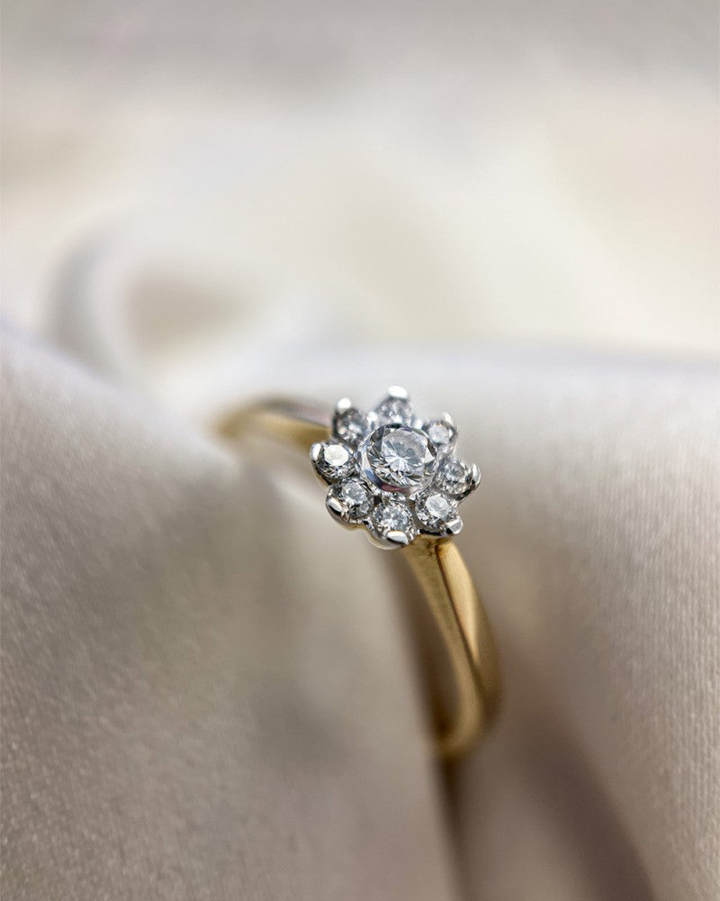 De Elegante Bicolor Ring met Briljant Diamanten - 0.21 crt van Excellent Jewelry, uitgevoerd in geel- en witgoud met een bloemvormig cluster van briljanten, is gepresenteerd op een zachte off-white stoffen achtergrond.