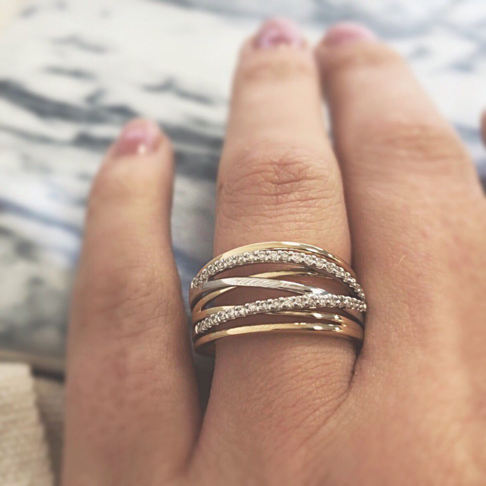 Een hand met roze nagellak showt de Excellent Jewelry Bicolor Gouden Crossover Ring met Diamant - 14 karaat, met kriskras door elkaar lopende banden, sommige bezet met diamanten, tegen een onscherpe achtergrond.