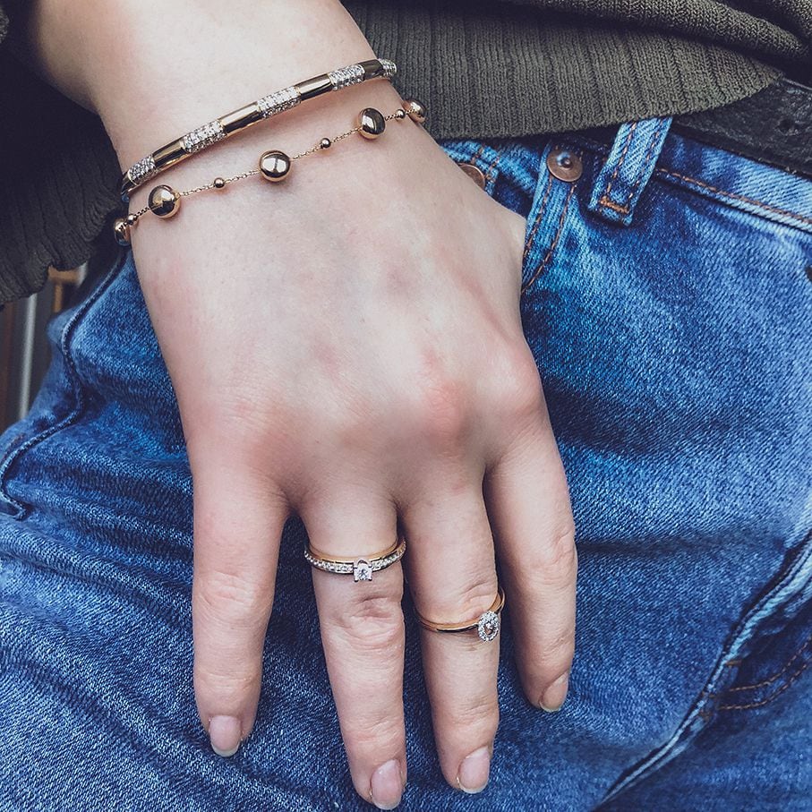 Een hand draagt de Excellent Jewelry Elegante Geelgouden Damesring met Witgouden Zetting en Briljant van 0,30 crt, gecombineerd met twee gouden armbanden, een blauwe spijkerbroek en een gedeeltelijk zichtbare groene trui.