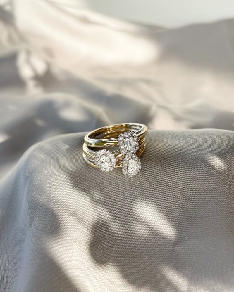 Een stapeltje van vier 14k gouden ringen, met Excellent Jewelry's 14k Bicolor Gold Ring met een smaragd geslepen diamant in een entourage zetting, getoond op zacht lichtgrijs satijn met gedempt licht en schaduw.