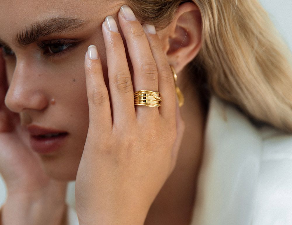 Een close-up van een lichtgetinte, blonde persoon die zijn gezicht aanraakt en de Excellent Jewelry Speelse Geelgouden Damesring-Brede Ring met Speelse Banen in 14 karaat goud en een gouden hoepel draagt, gekleed in lichte kleuren.