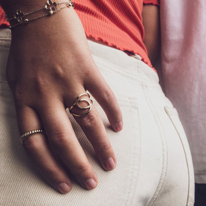 Een close-up van een hand met de Geelgouden Damesring - 14 Karaat Goud met Strak Design van Excellent Jewelry en een armband, rustend op de achterzak van een lichte spijkerbroek. De persoon draagt een koraalkleurig topje met textuur.