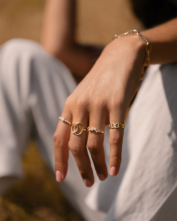 Een close-up van een hand met een brede ring en verschillende gouden armbanden, met de Excellent Jewelry Geelgouden Damesring met Speels Design - 14 Karaat Goud. De persoon in de witte broek zit buiten in natuurlijk licht.