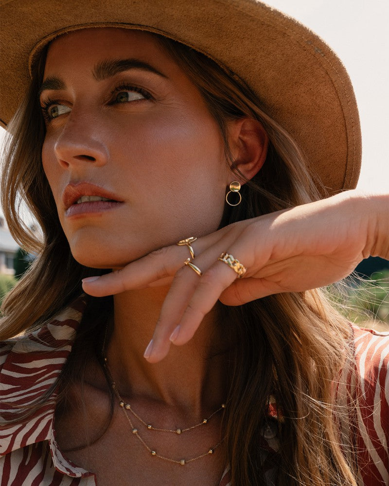 Buiten draagt een vrouw met lang haar en subtiele make-up een bruine hoed, shirt met patroon en een Excellent Jewelry 14 karaat geelgouden damesring (6 mm ras), haar hand opgeheven om het eenvoudige ontwerp te laten zien.