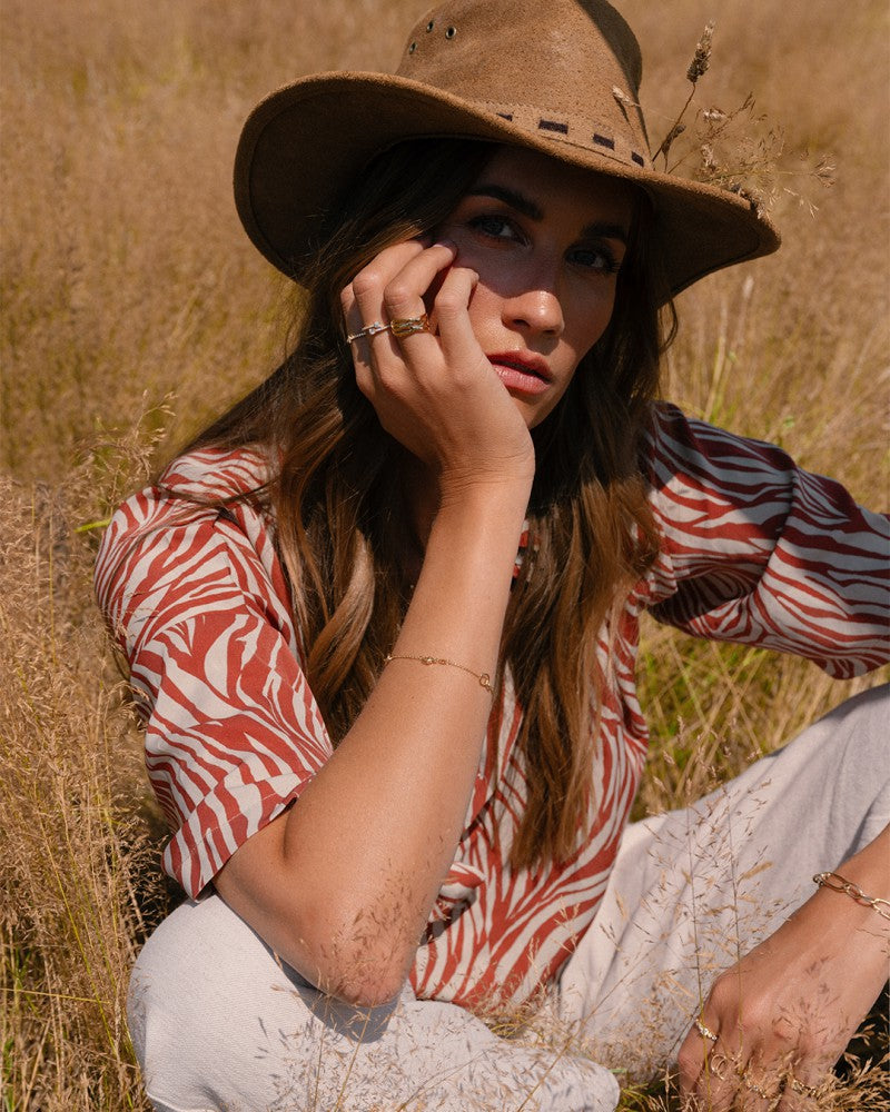 Een vrouw met een bruine hoed, een rood-wit shirt met patroon en het Excellent Sieraad Geelgouden Damesring - Eenvoudig en Elegant in 14 Karaat Goud zit in hoog gras, laat haar kin op haar hand rusten en kijkt peinzend naar de camera.
