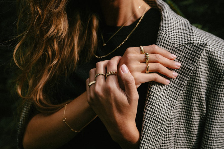 Een vrouw met golvend haar, die een geruite blazer en een zwart topje draagt, showt verschillende gouden ringen - waaronder de Excellent Jewelry Geelgouden Gedraaide Bandring (14kt goud, 4,3 mm breed)- terwijl ze haar handen zachtjes tegen haar borst houdt.