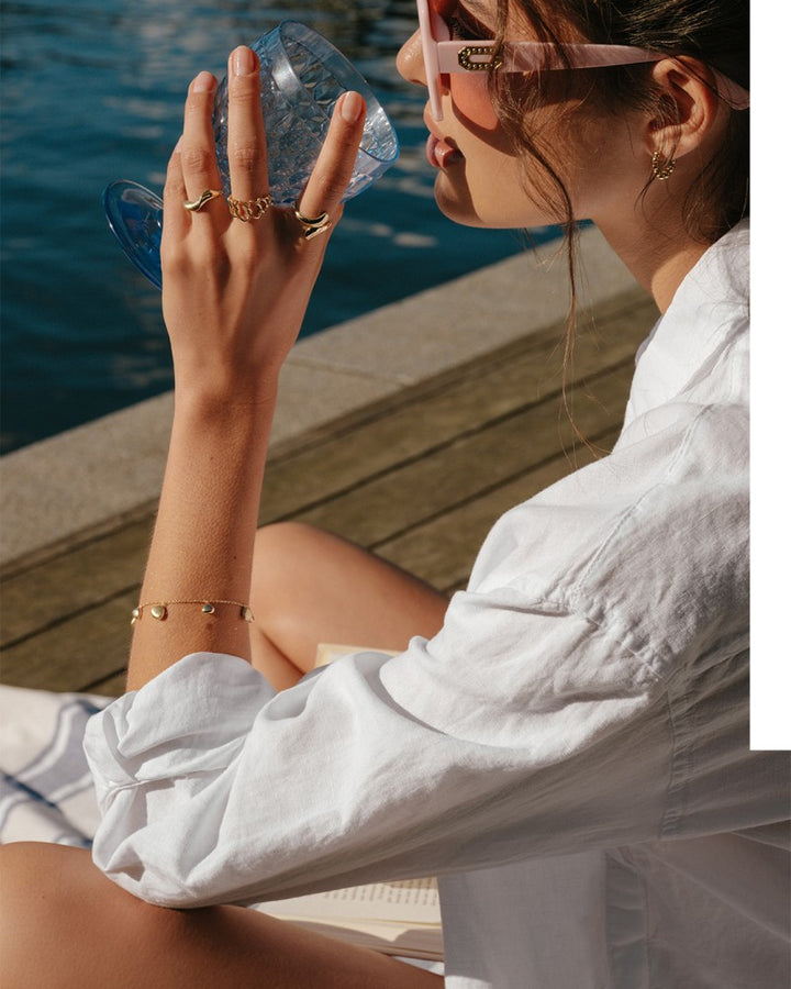 Een vrouw in een wit shirt en een zonnebril ontspant op een zonovergoten houten dek aan het water en draagt de Geelgouden Ring met Organische, Speelse Twist - Een Vleugje Natuur in Jouw Stijl van Excellent Jewelry, samen met andere gouden sieraden.