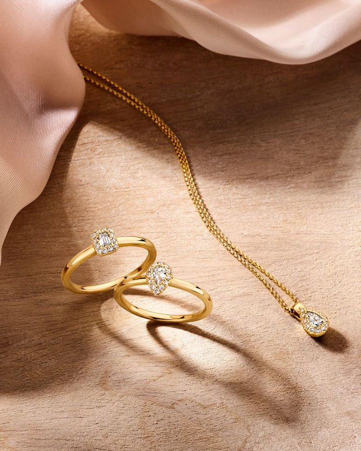 Twee verfijnde geelgouden ringen met baguette diamant 0.24 crt van Excellent Jewelry en een gouden ketting met peervormige diamant hanger, stijlvol gepresenteerd op hout naast zacht lichtroze stof.