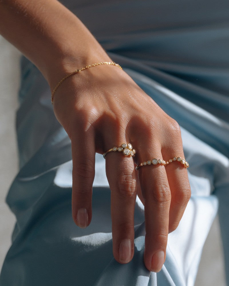 Een close-up van een hand die Excellent Jewelry's Elegante gouden ring met 0,32ct diamanten in een 'Bubbles' design draagt, gemaakt in 14k geel goud, rust op lichtblauwe satijnen stof naast delicate gouden ringen en een dunne armband.