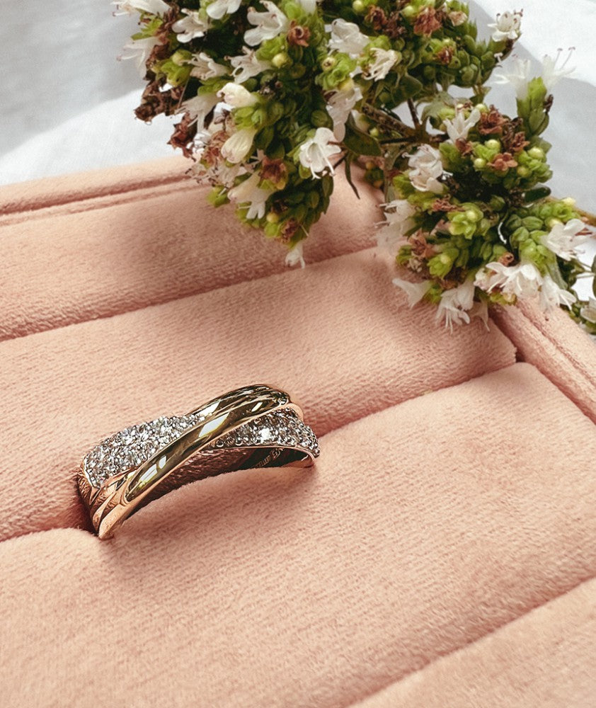 De Bicolor Geel- en Witgouden Crossover Ring met Diamanten van Excellent Jewelry (14k goud, 0.40ct diamanten) roest op een blushroze fluwelen ringdoosje, met een klein boeket witte bloemen ernaast.